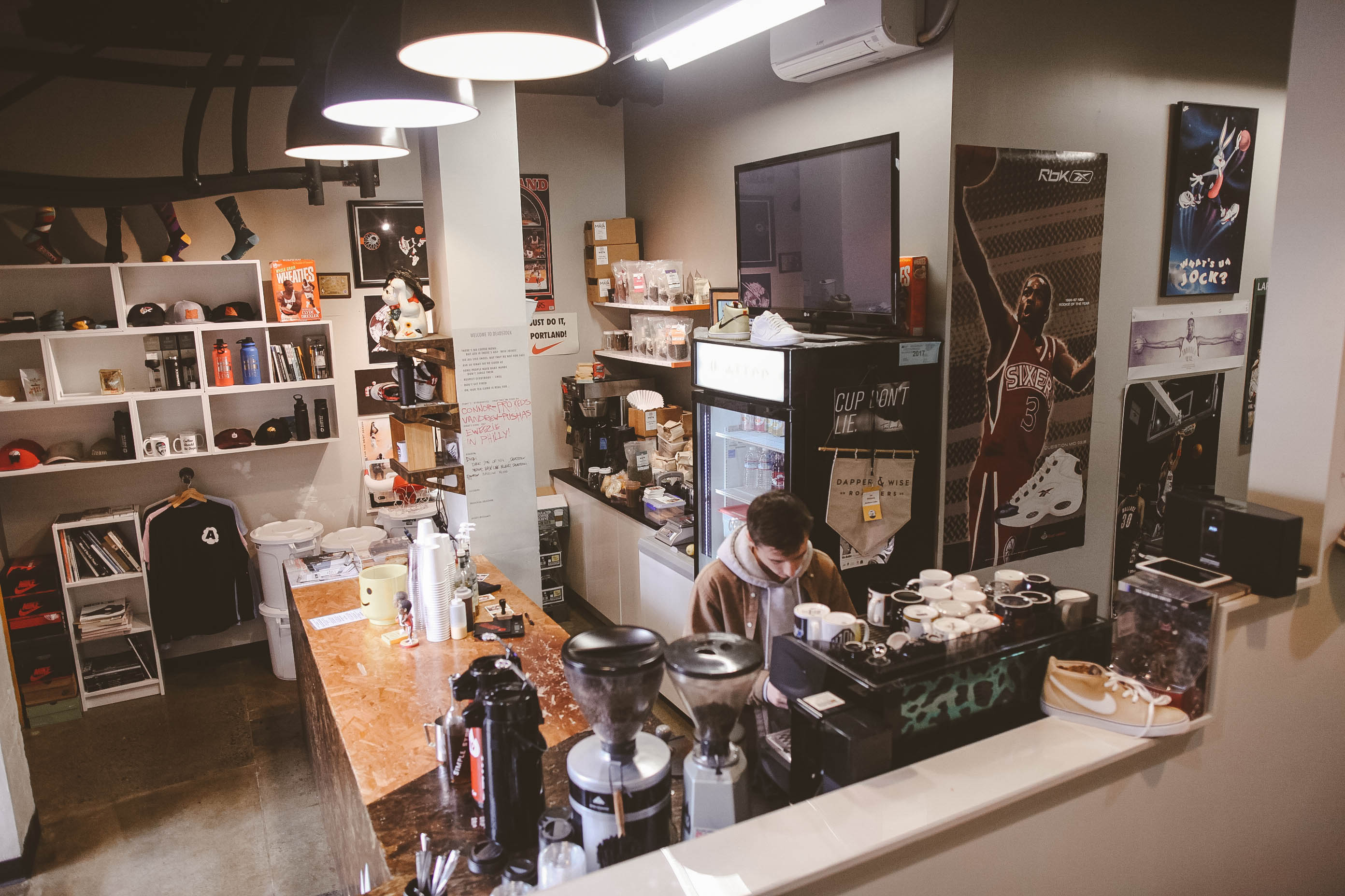 A man in brown jacket and gray hoodie works behind an espresso machine in a coffee sports cafe, as a basketball Sixers Reebok poster hangs on the wall.