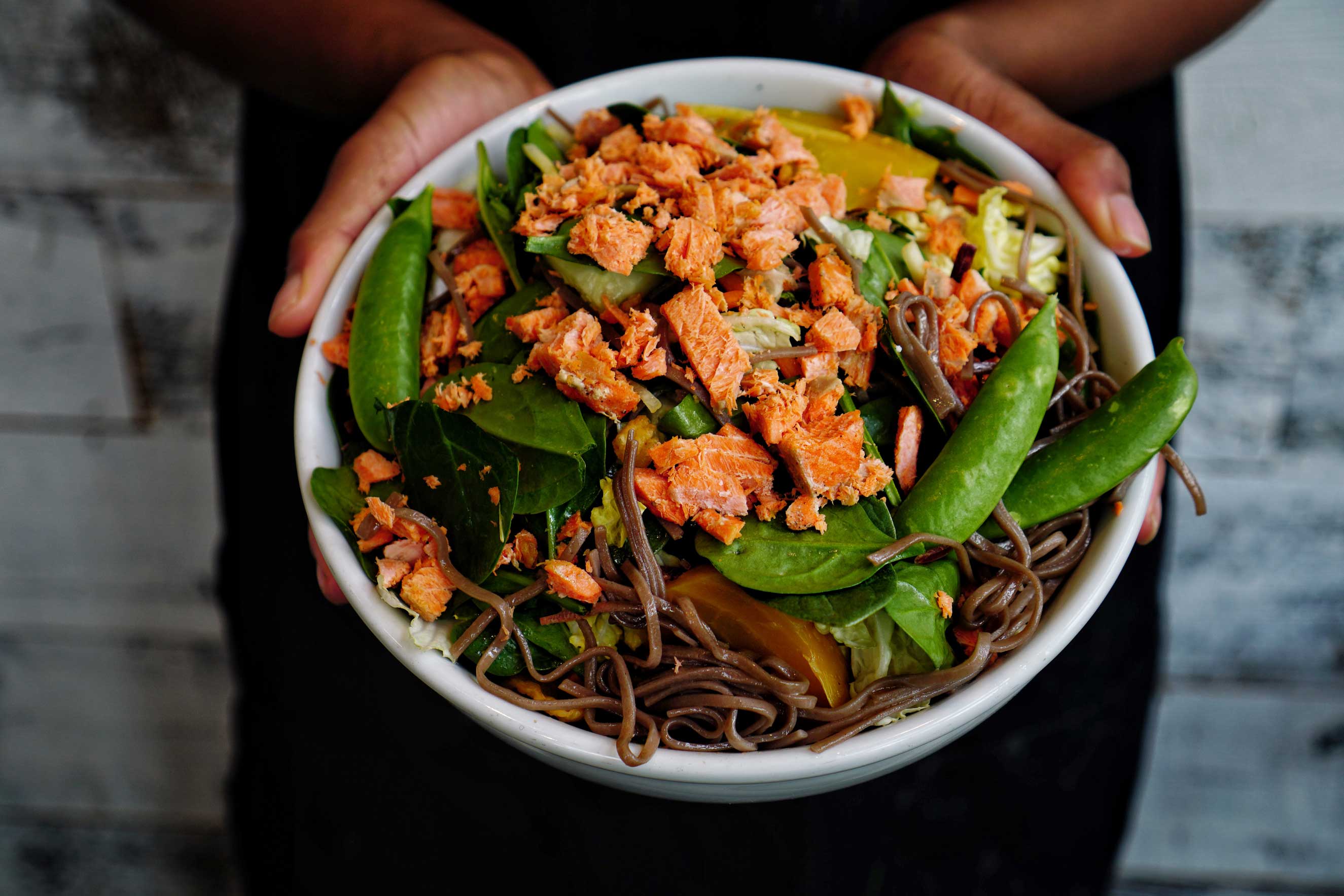Two hands holding a white bowl of brown noodles, spinach leaves and salmon.