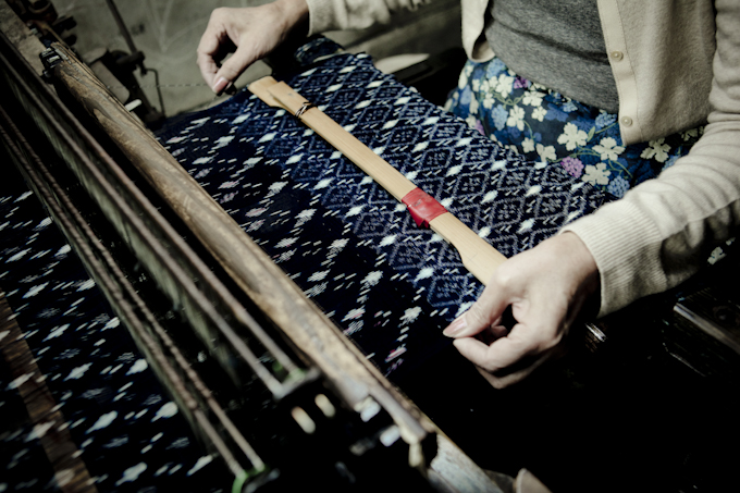 A woman (faced not pictured) works on making a patterned material on a wooden machine.