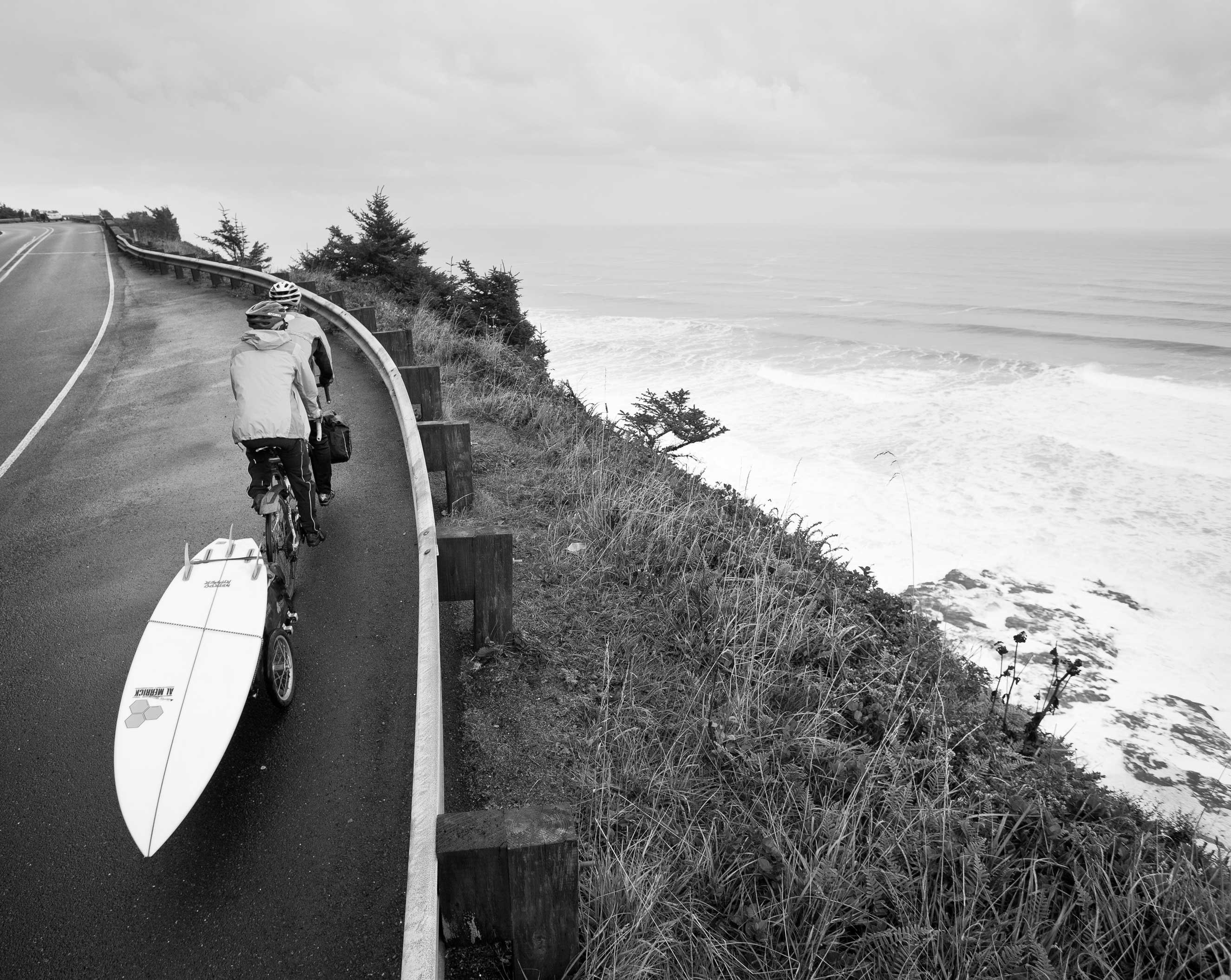 Two bicyclists cycle along the road with helmets and jackets on, towing a long a surfboard. Site of the ocean off to the right.