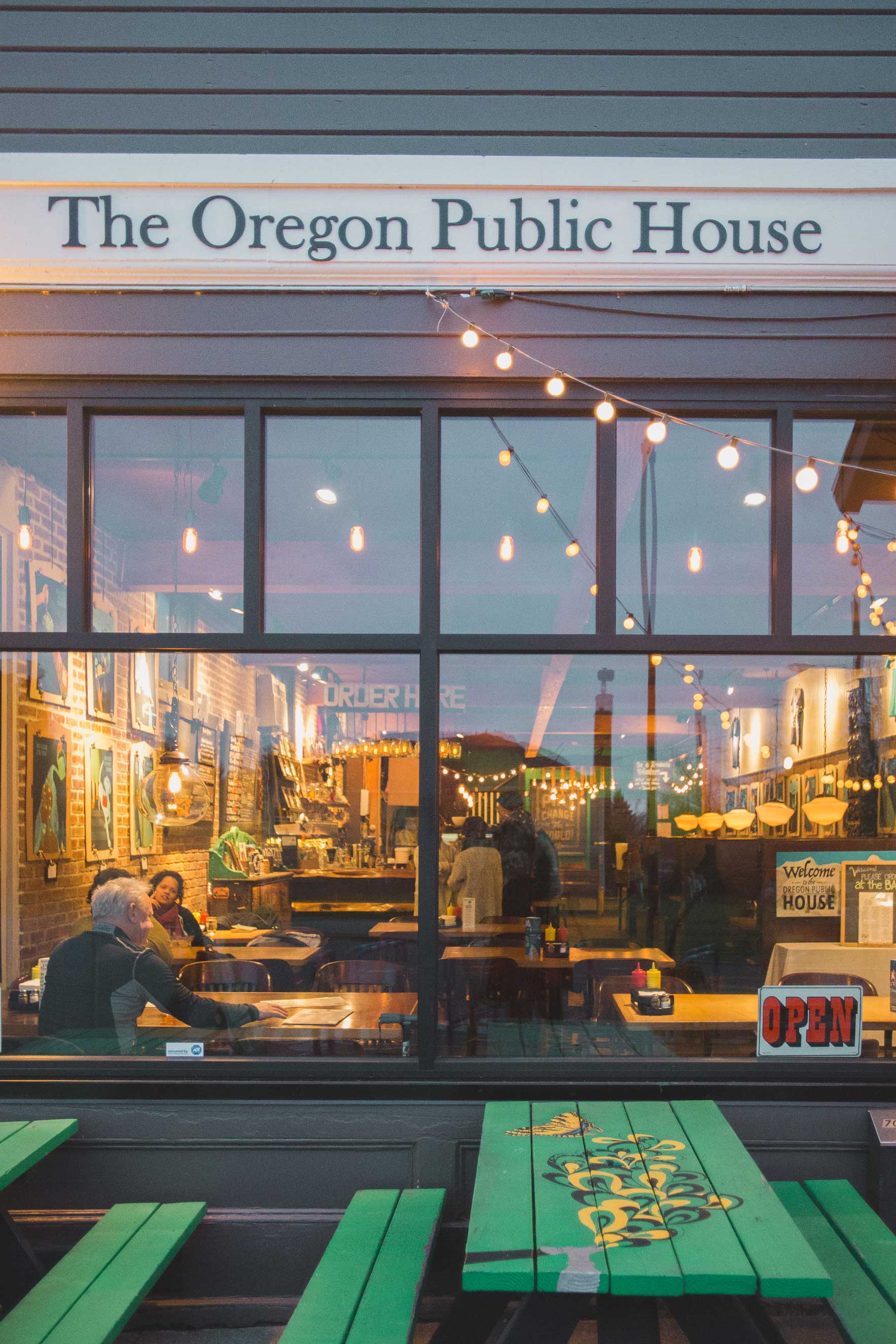 The exterior of The Oregon Public House, with open sign in the window and green painted wooden tables pushed up against the window.