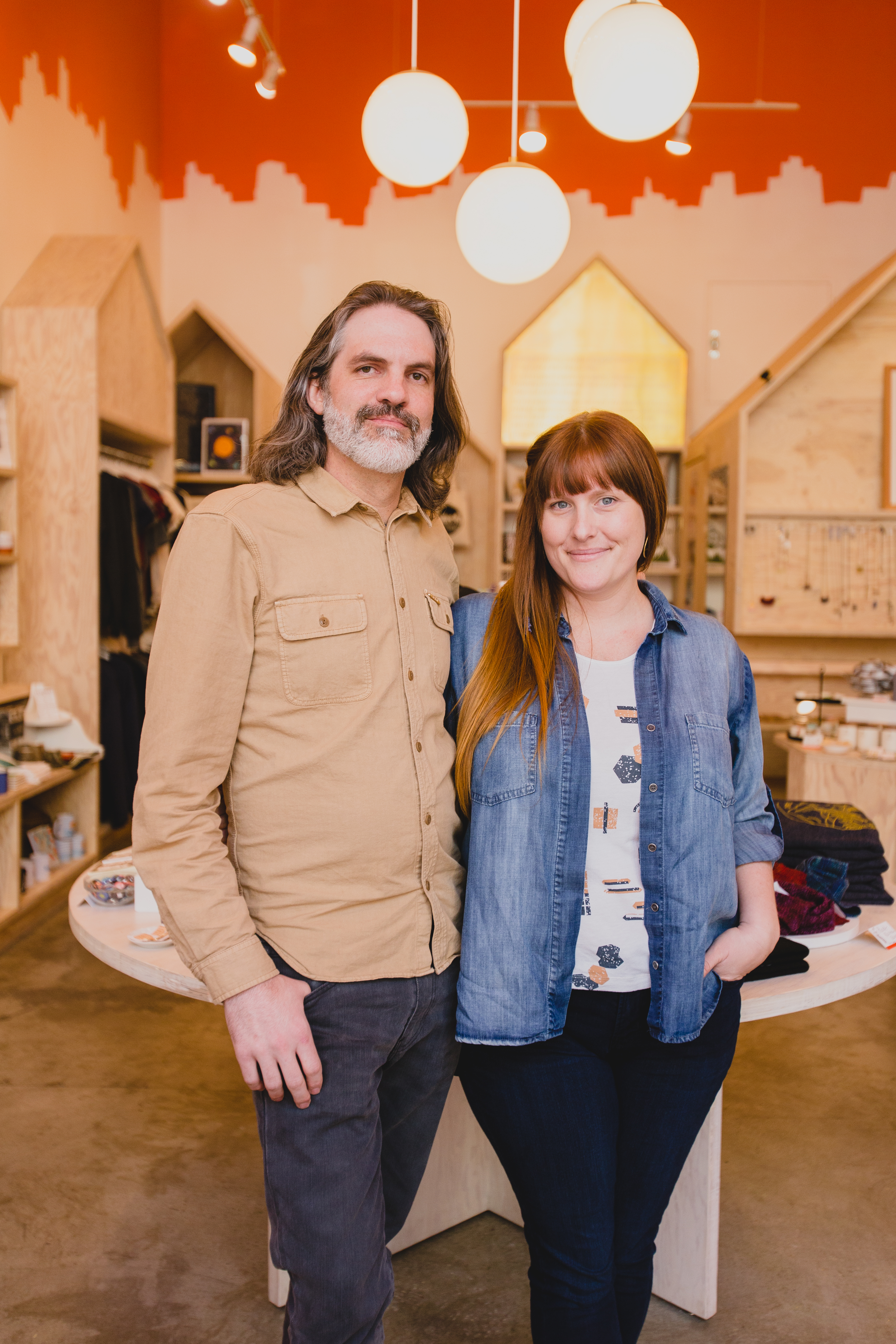 Brianne—chambray top layered over patterned shirt w/dark denim—and husband Jared Mees—tan long-sleeved button up shirt and gray denim, smile for the camera.