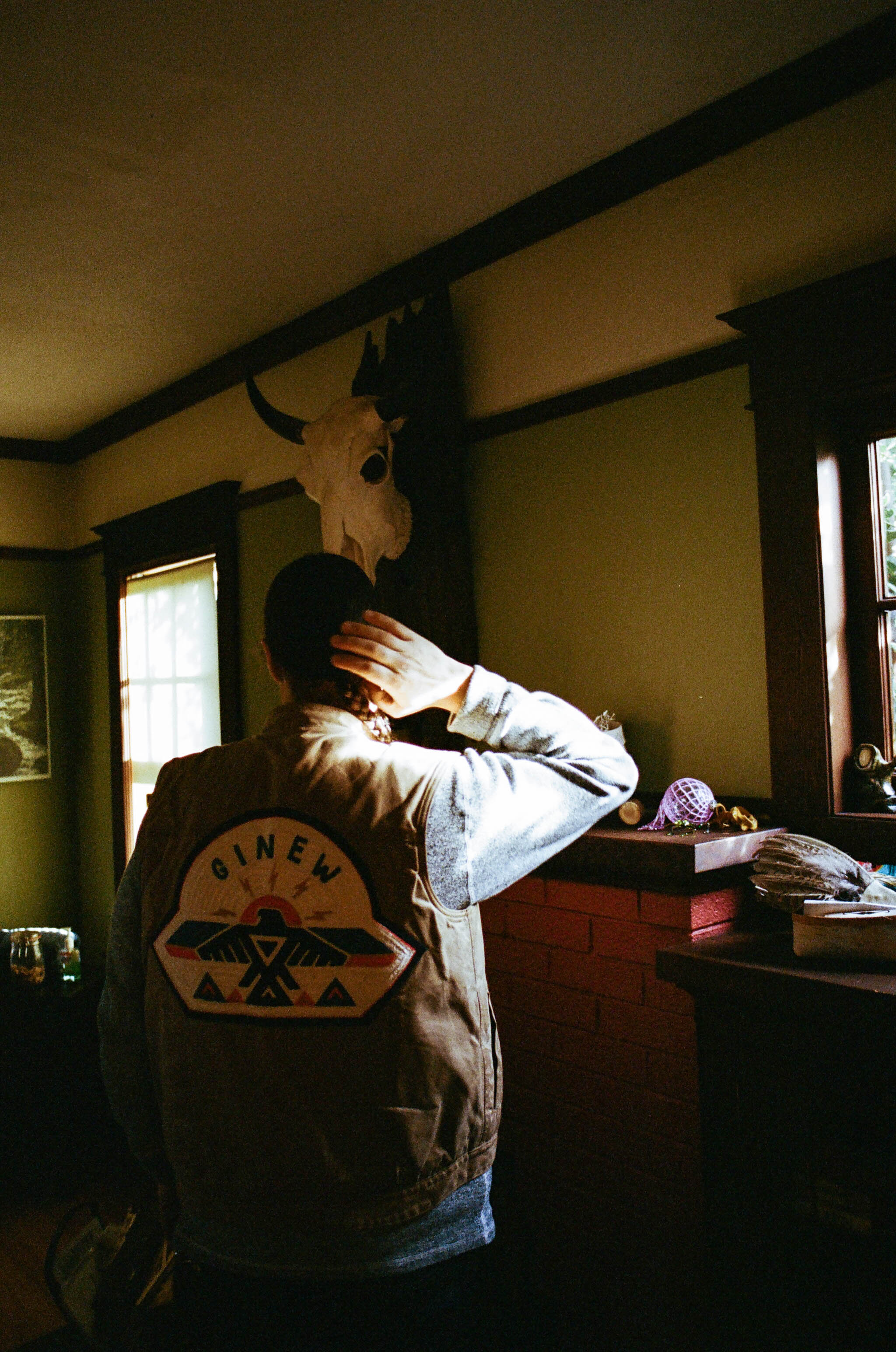 A man stands with his back to the camera showcasing his brown leather Ginew vest layered over a long-sleeve shirt. A skull of an antelope hangs on the wall behind.