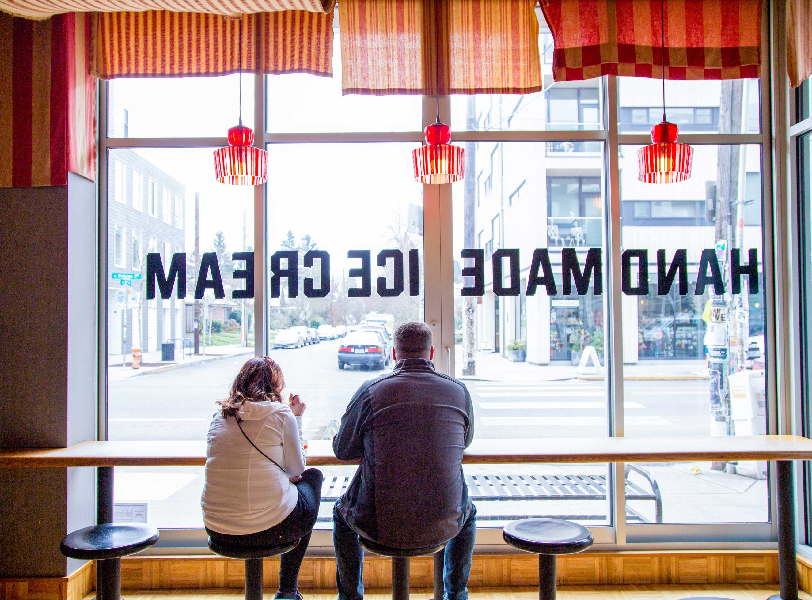 Two people—female in white jacket and male in denim—sit looking out the window of a handmade ice cream shop.