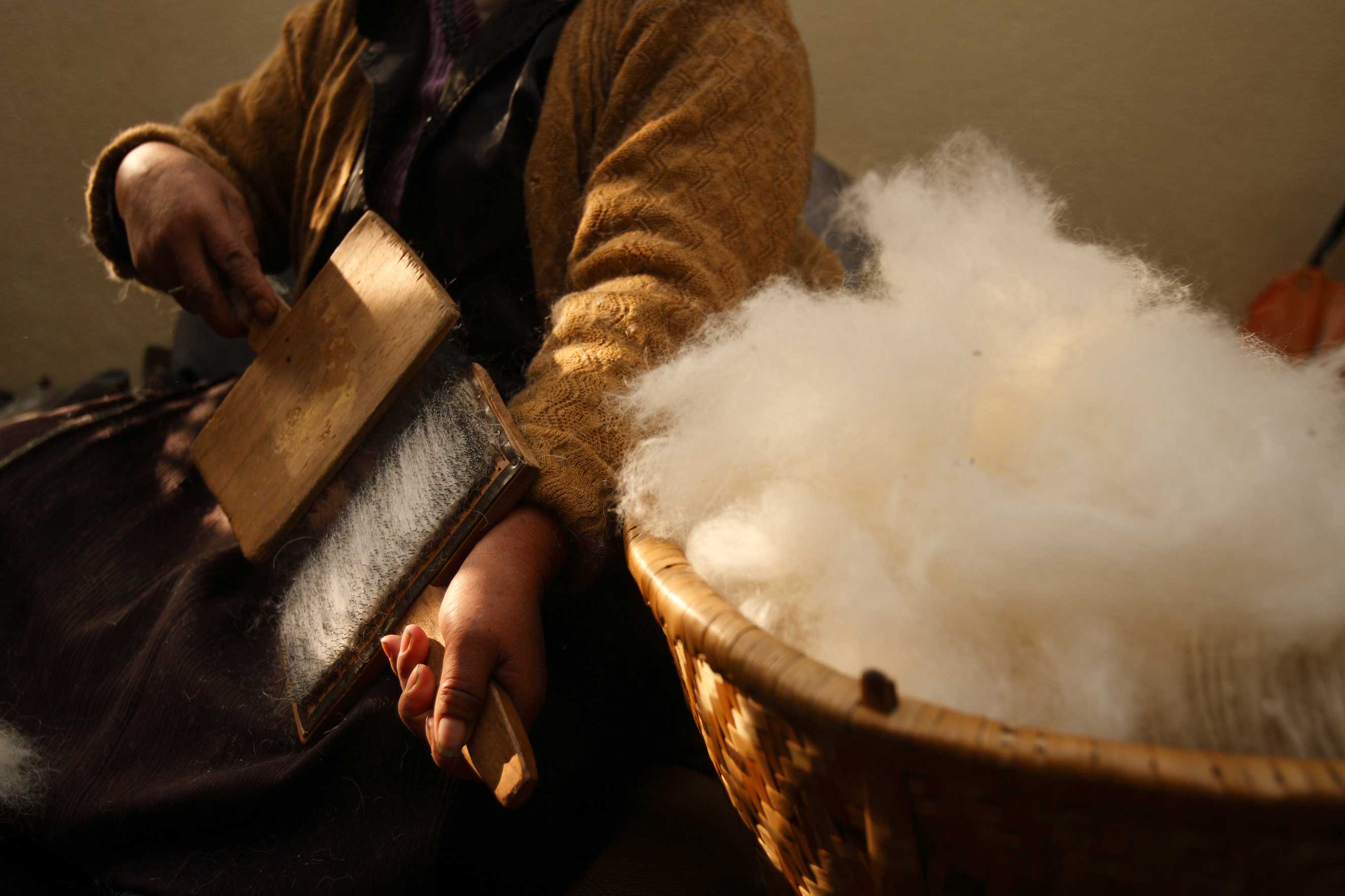 A lady (face not pictured) in a yellow knitted cardigan, brushes white wool between two brushes, with a basket of white wool sitting in foreground.