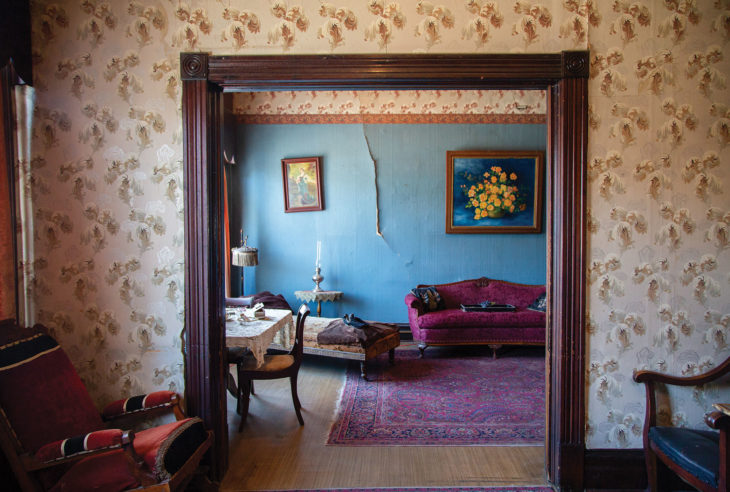 Faint feather patterned wallpaper lines the room walls with wooden beam entryway. A crack in turquoise wallpaper in the background with maroon velvet couch and oil flower paintings.