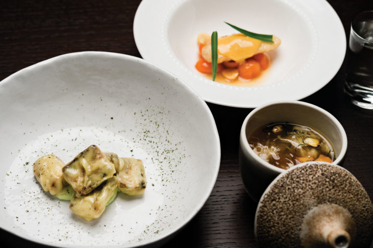 Three Japanese dishes, two on white plates and one in a white bowl with lid leaning against it.