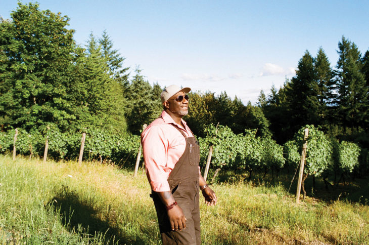 A black man with tan hat, sunglasses, salmon colored long-sleeve shirt, brown overalls, and red beaded bracelet stands in the foreground of a vineyard.