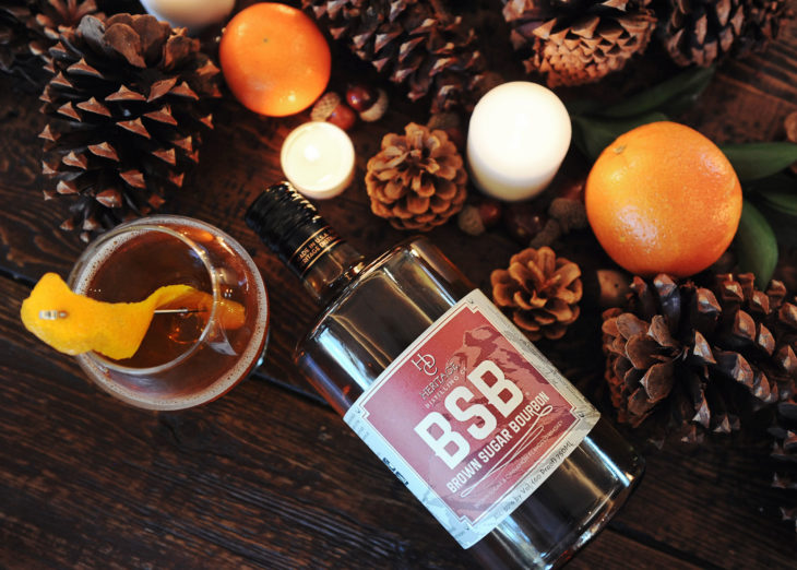 Heritage Distilling Co.’s Brown Sugar Burbon, Alc 30% by Vol (60 Proof) 750ML—surrounded by white lit candles, oranges, and pinecones w/drink.
