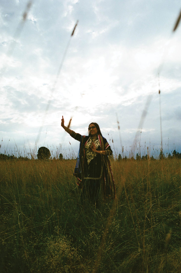 Woman in dark colored clothing and shaw dancing in a long-grass field.