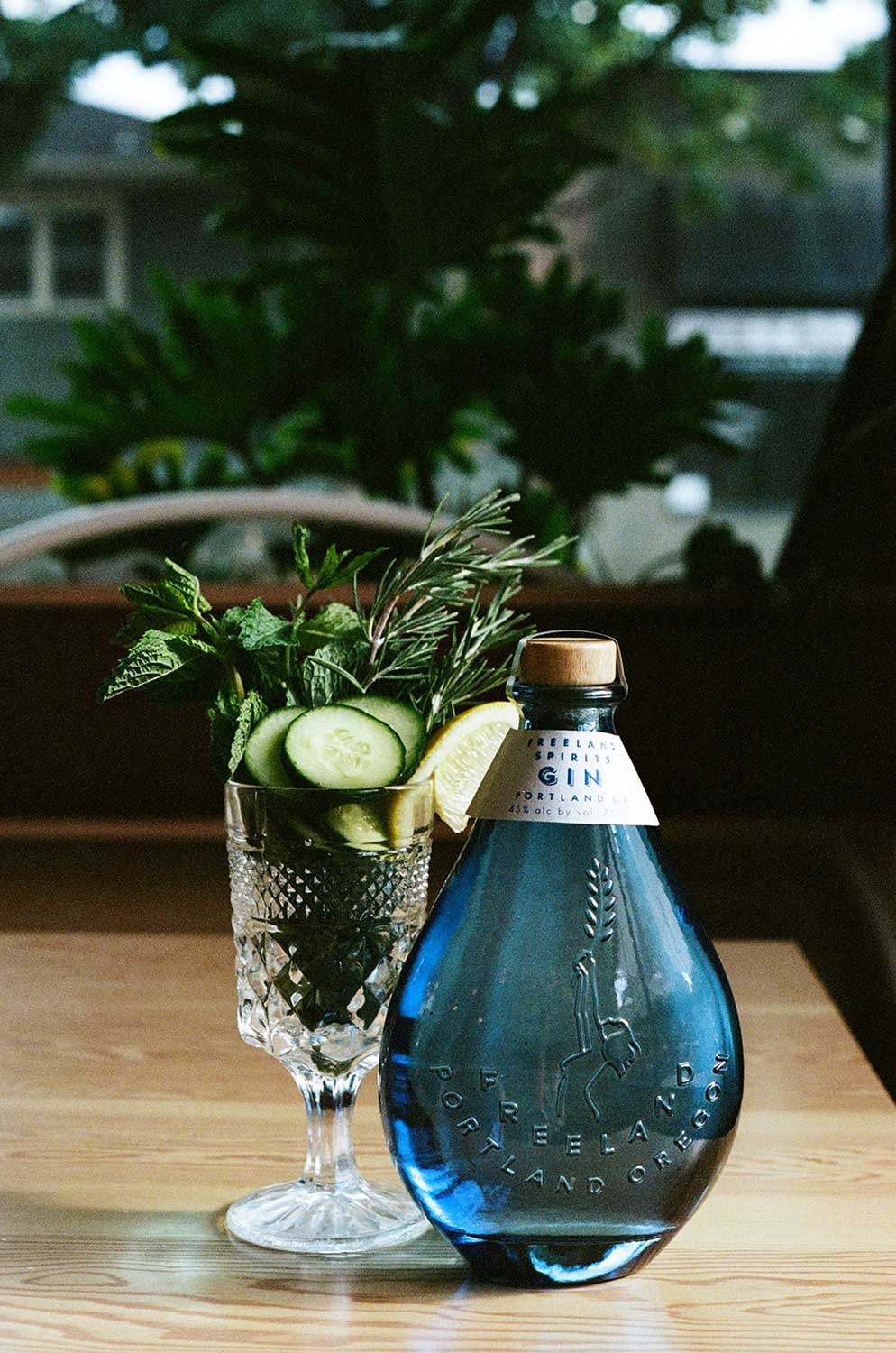 An elegant cup filled with herbs, cucumbers, and sliced lemons next to a blue bottle of gin