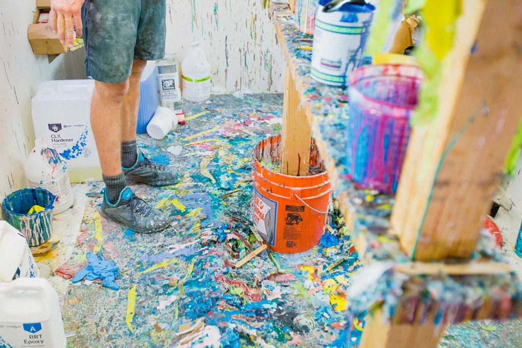 A person standing on a paint splattered workshop floor with buckets of paint surrounding him